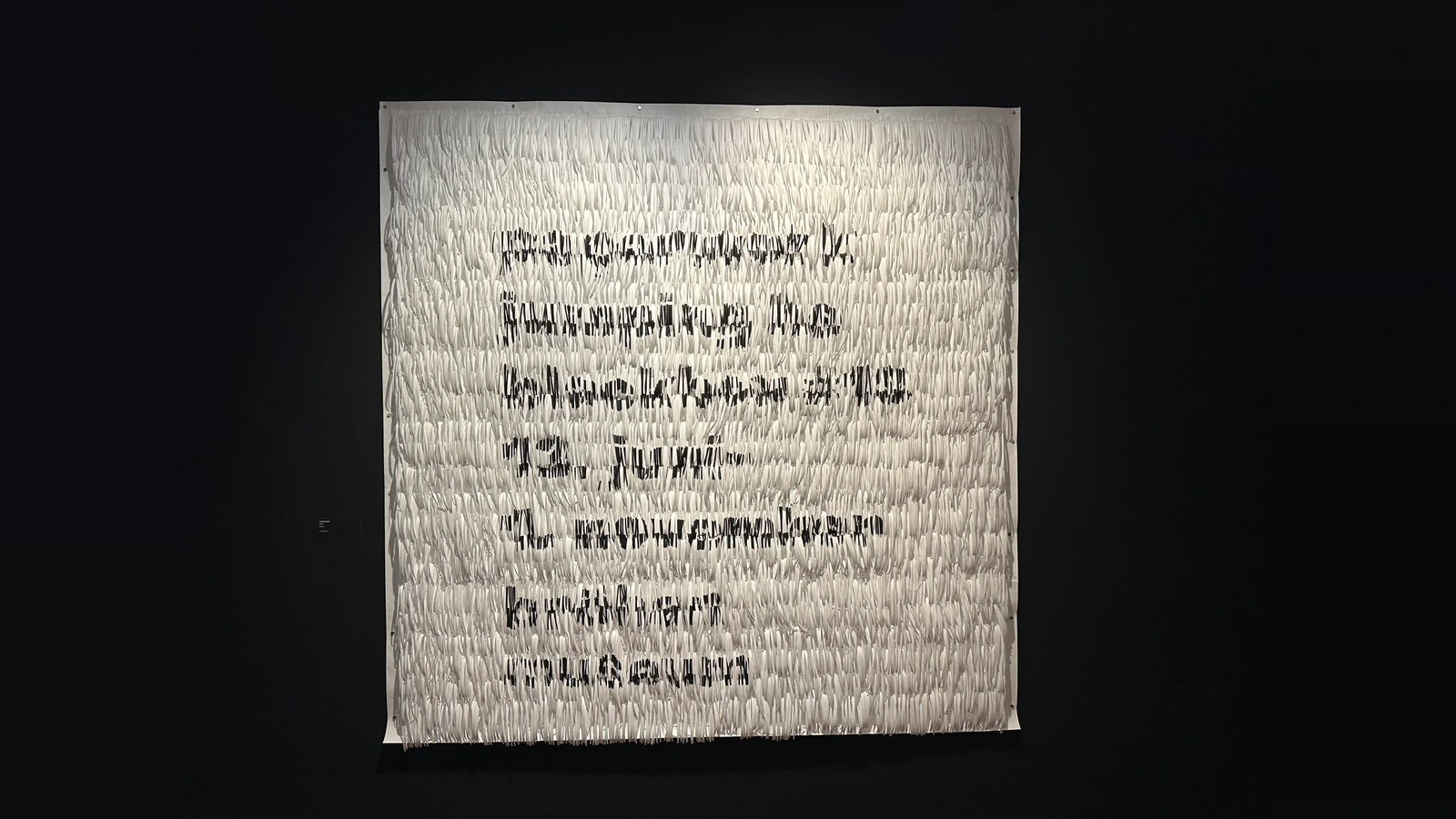 Helle Papierarbeit mit zahlreichen schmalen Papierpartien und schwarzen Schriftfragmenten vor dunkler Wand